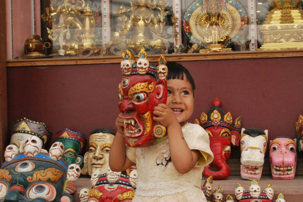 Young girl holds a vibrant cultural mask in a souvenir shop in Kathmandu, Nepal, surrounded by colourful sculptures.